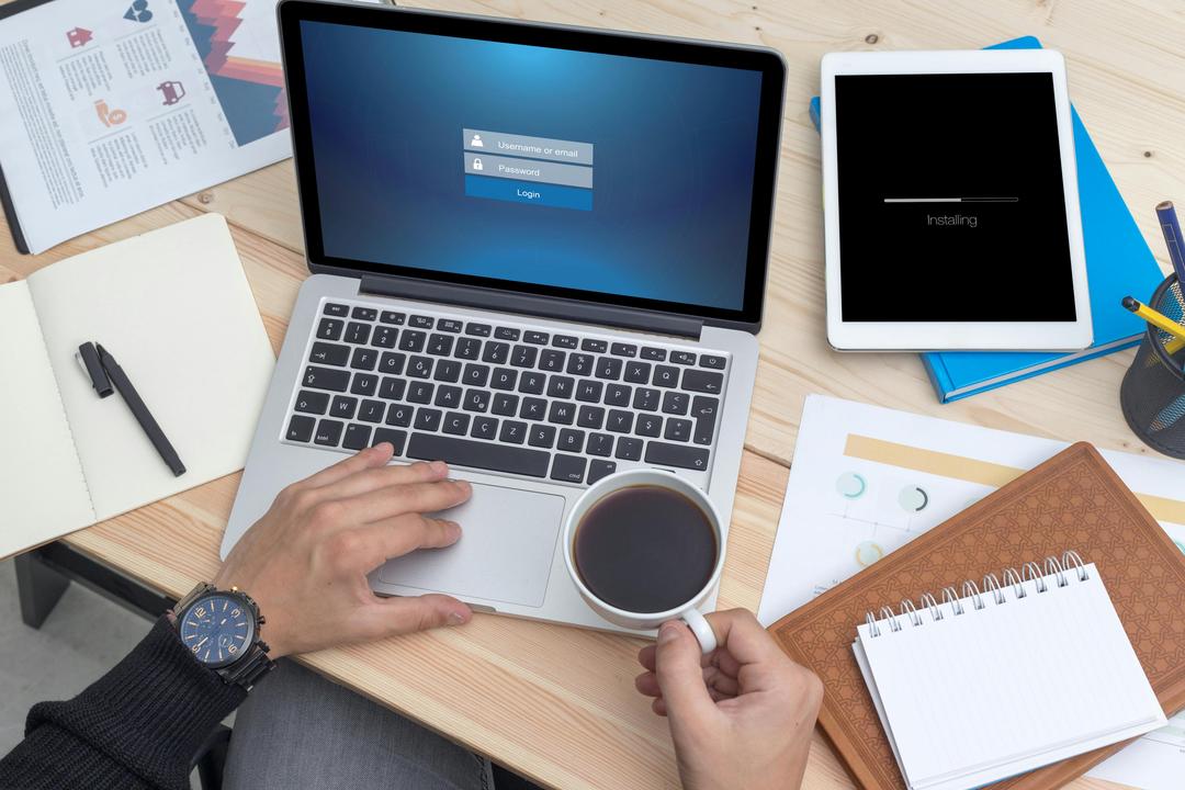 a table with a laptop, notebooks, and iPad with a person holding a coffee